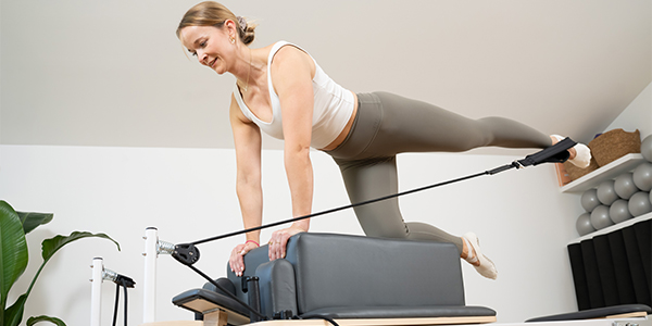 Eine Person trainiert auf dem Taurus Reformer Pilates Pro und kniet dabei auf der Box, ein Fuß ist in der Schlaufe.