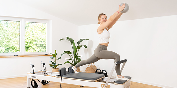 Eine Person steht im Ausfallschritt auf dem Reformer und bewegt mit einem Bein den Schlitten nach hinten. In den Händen hält sie einen Ball nach oben. 
