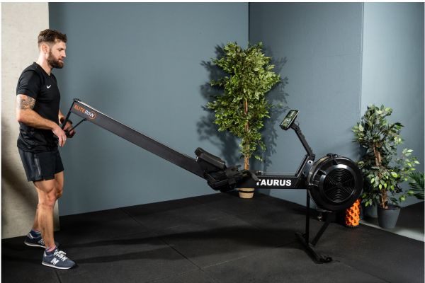 Eine Person in Sportbekleidung hebt den Taurus Elite Rower am hinteren Standfuß an.