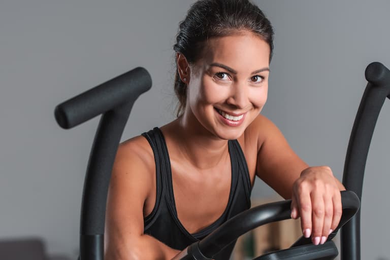 Nahaufnahme einer Person, die auf einem Crosstrainer steht und in die Kamera l&auml;chelt. Mit dem linken Arm st&uuml;tzt sie sich locker auf dem mittleren Teil des Crosstrainers ab. Der Hintergrund ist gr&auml;ulich und unscharf.
