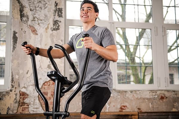 Person trainiert im Wohnraum auf einem Crosstrainer. Die Person h&auml;lt die Trainingsarme fest und l&auml;chelt. Im Hintergrund eine Fensterfront und eine Wand.