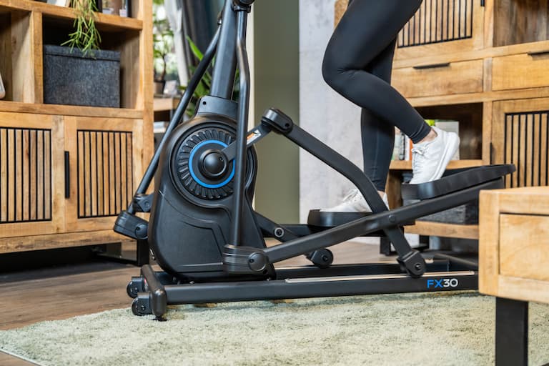 Nahaufnahme des unteren Teils eines Crosstrainers, auf dem eine Person trainiert. Der Crosstrainer steht in einem Wohnzimmer.