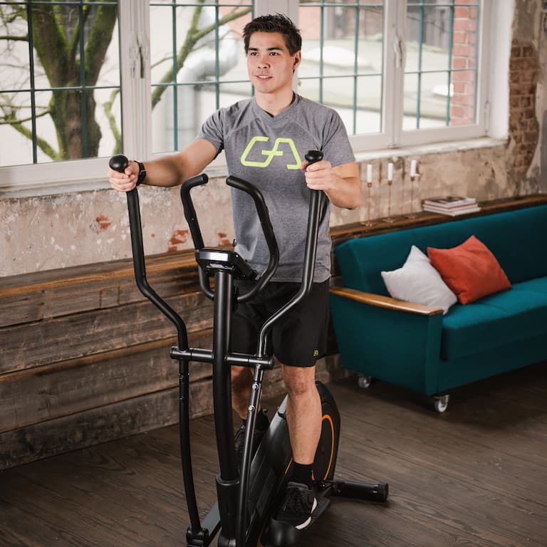 Eine Person trainiert auf einem Crosstrainer in wohnlichem industriellen Ambiente vor einer Fensterfront. 