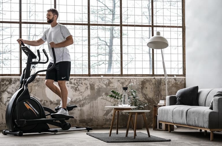 Eine Person trainiert in Sportbekleidung in wohnlicher Umgebung auf einem Taurus Crosstrainer. Im Hintergrund ist eine gro&szlig;e Fensterfront, eine Couch und eine Lampe.