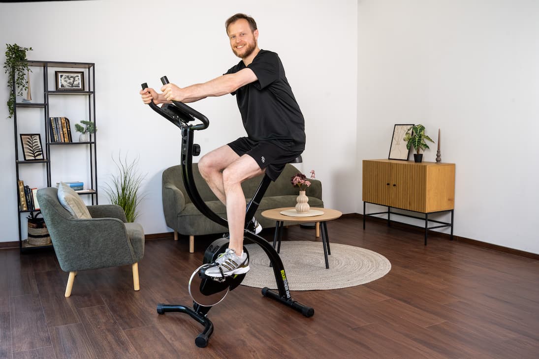 Person trainiert auf einem Heimtrainer. Das Trainingsgerät ist in einem Wohnzimmer platziert. Die Person trägt sportliche Kleidung und lächelt.