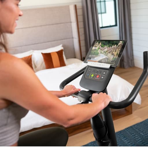 Blick über die Schulter auf Lenker und Konsole eines Heimtrainers, zudem ein Tablet auf der Tablet-Halterung. Eine Person trainiert gerade auf dem Heimtrainer und lächelt. Sie nutzt eine Streckenapp. Im Hintergrund ist ein Bett zu sehen.