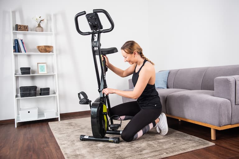 Eine Person hockt in sportlicher Kleidung neben einem aufgestellten Duke Fitness Heimtrainer XB30 und hält mit der einen Hand die Sattelstange und mit der anderen Hand eine Stellschraube.