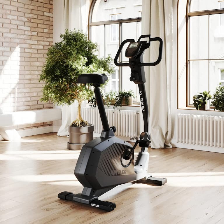 Produktbild des Heimtrainers Kettler Giro M, welches schräg von der Seite gezeigt wird. Der Heimtrainer steht in einem Wohnzimmer.
