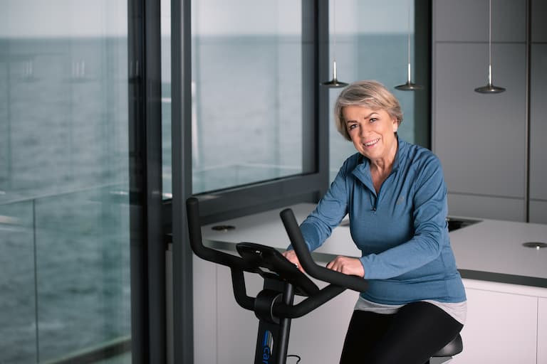 Ältere Person sitzt auf einem Heimtrainer in wohnlicher Umgebung. Sie trägt Sportkleidung und lächelt. Im Hintergrund sind große Fensterfronten, dahinter ist das Meer zu sehen.