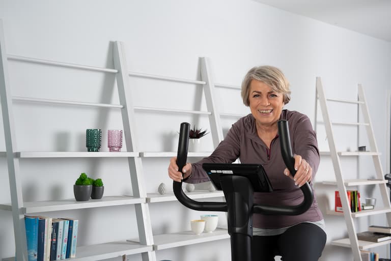 Eine Person sitzt in einer wohnlichen Umgebung in Sportbekleidung auf einem Heimtrainer. Im Hintergrund ist ein langes Regal zu sehen.