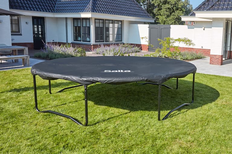 Ein Trampolin mit Wetterschutzplane steht in einem Garten auf gr&uuml;nem Rasen. Im Hintergrund ist links eine Terrasse zu sehen und der ganze Hintergrund wird von dem Haus bedeckt. Zwischen Haus und Trampolin sind Beete mit Lavendel und Gr&uuml;npflanzen, die Sonne scheint, der Himmel ist blau.