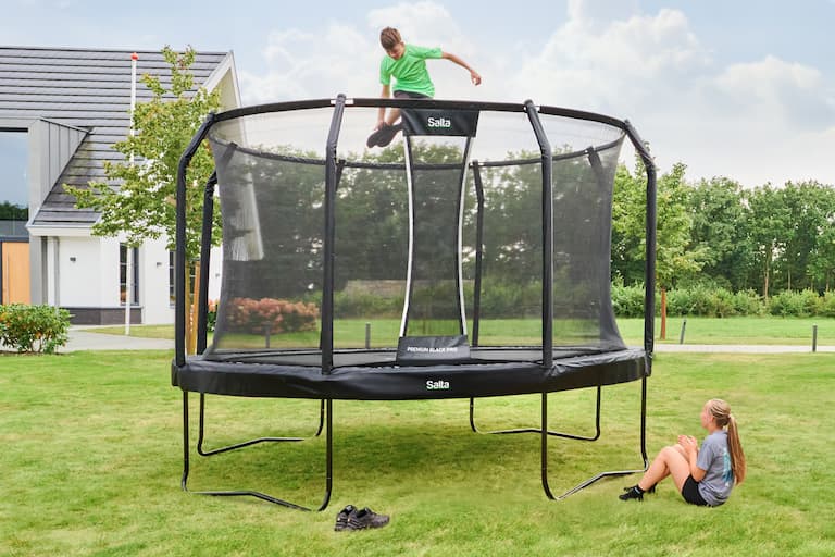 Ein Kind springt auf einem runden Trampolin mit Standbeinen im Garten, vor dem Trampolin sitzt ein weiteres Kind und sieht dem Jungen zu.