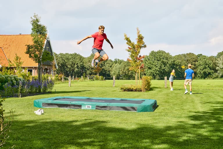 Ein Kind springt auf einem eckigen Bodentrampolin im Garten, im Hintergrund spielen zwei weitere Kinder mit einer Frisbee.