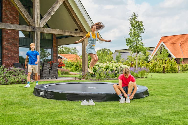 Ein M&auml;dchen springt auf einem runden FlatGround Trampolin im Garten. Auf dem vorderen Rand des Trampolins sitzt ein Junge und bindet sich die Schuhe. Im Hintergrund ist ein Haus zu sehen, der Garten ist gr&uuml;n und verschiedene Pflanzen und Blumen bl&uuml;hen. Links neben dem Trampolin ist ein Junge zu sehen, der am Trampolin vorbeil&auml;uft und das M&auml;dchen ansieht.