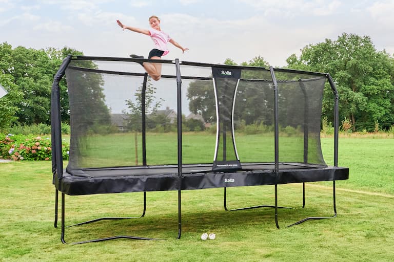 Ein M&auml;dchen springt auf einem rechteckigen Trampolin mit Sicherheitsnetz hoch in die Luft und nimmt oben eine Pose ein. Das Trampolin steht in einem Garten, im Hintergrund sind gr&uuml;ne B&auml;ume zu sehen. 