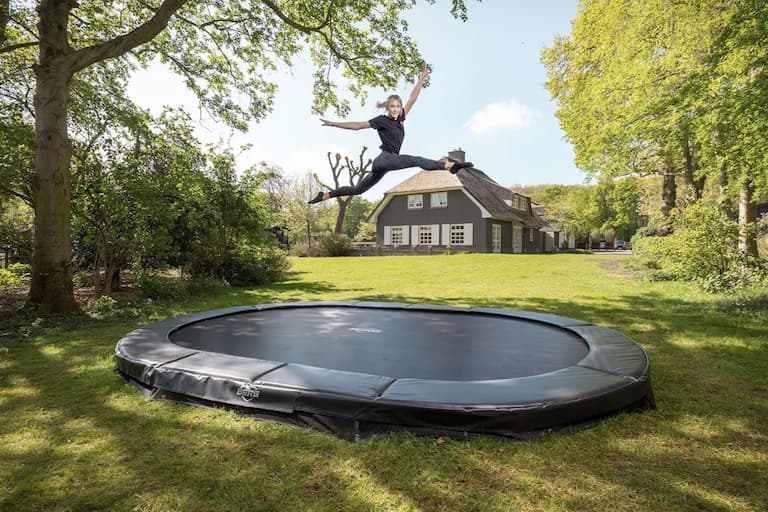 Ein Ovales Trampolin, welches im Boden eingelassen ist, steht zwischen gr&uuml;nen B&auml;umen in einem Garten. Im Hintergrund ist ein Reetdachhaus zu sehen mit grauer Fassade und wei&szlig;en Fenstern. Auf dem Trampolin springt ein M&auml;dchen hoch in die Luft und macht oben einen Spagat.