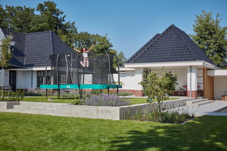 Auf dem Bild ist ein Garten zu sehen, im Hintergrund ein Haus. Der Garten hat mehrere Ebenen, vor dem Haus auf der erh&ouml;hten Ebene des Gartens steht ein rechteckiges Trampolin mit Sicherheitsnetz, auf dem ein M&auml;dchen springt.