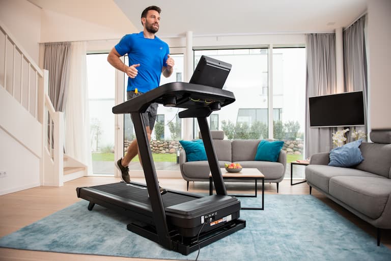 Person läuft auf dem cardiostrong Laufband TX70. Das Laufband befindet sich in einem Wohnzimmer. Im Hintergrund ist eine Fensterfront, Couches sowie eine Treppe zu sehen.
