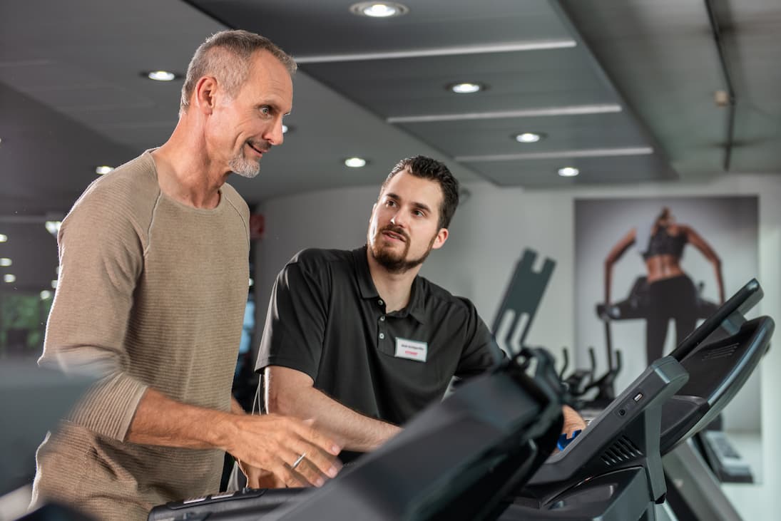 Eine Person (Kunde) steht auf einem Laufband und blickt auf die Konsole des Laufbands. Das Laufband befindet sich in einer Fitshop-Filiale. Neben dem Kunden steht ein Fitshop-Mitarbeiter, blickt den Kunden freundlich an und spricht.
