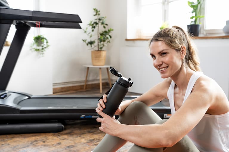 Im Vordergrund sitzt eine Person in Sportkleidung mit einer sportlichen Trinkflasche in der Hand auf dem Boden innerhalb eines Wohnzimmers. Sie lächelt. Im Hintergrund ist ein Laufband zu sehen, sowie eine Fensterfront.