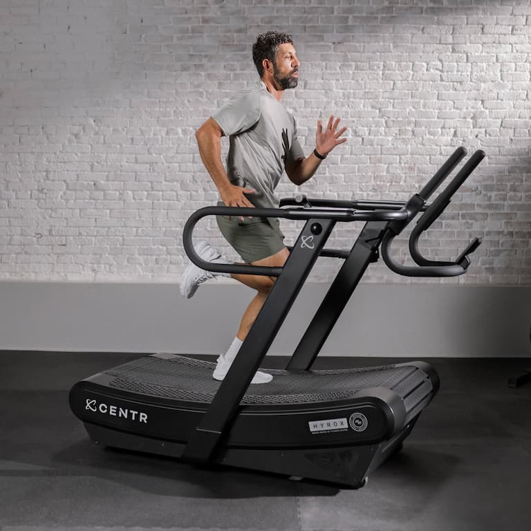 Person trainiert auf dem Centr x Hyrox Laufband Perform Tread. Das Laufband steht auf einem dunklen Untergrund vor einer weiß gestrichenen Wand, auf welcher das Mauerwerk durchscheint.
