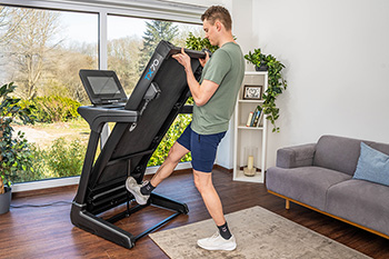 Eine Person in sportlicher Kleidung steht auf dem linken Bein und drückt mit dem rechten Fuß gegen den Klappmechanismus eines Laufbands, um es hoch- oder runterzuklappen. Die Person hält das Laufdeck am Ende fest, welches sich auf Höhe ihrer Schultern befindet. Das Laufband steht in wohnlichem Ambiente vor einer Fensterfront. 