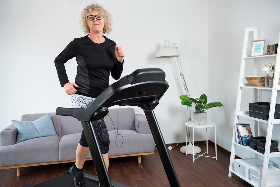 Person läuft auf einem Laufband. Das Laufband steht in einem Wohnzimmer. Im Hintergrund ein Sofa, ein Regal und weitere Einrichtungsgegenstände.
