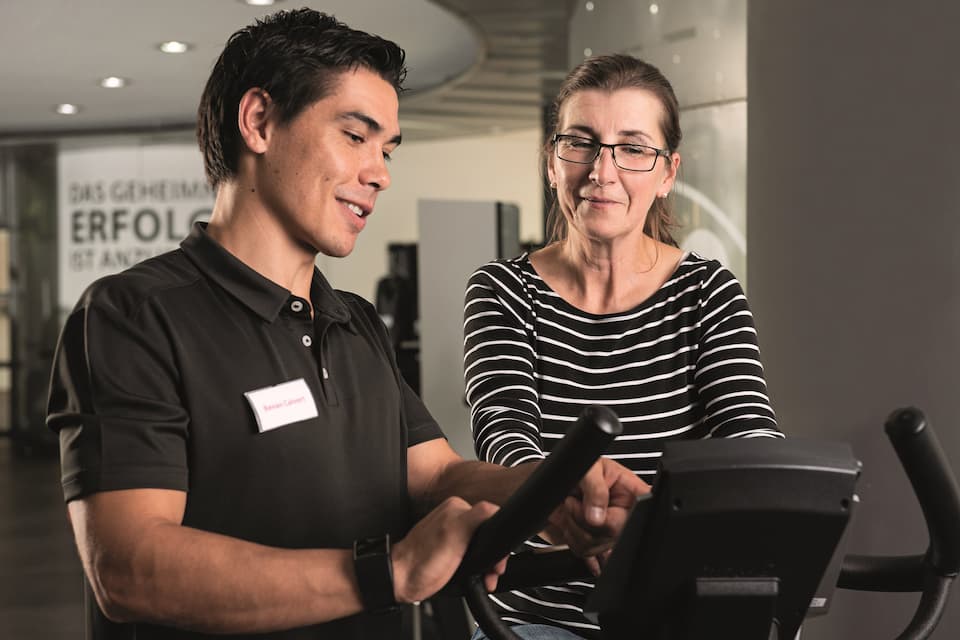 Eine Person wird von einem Fitshop-Mitarbeiter in einer Fitshop-Filiale beraten. Der Mitarbeiter zeigt der Kundin etwas an der Konsole eines Trainingsgeräts.