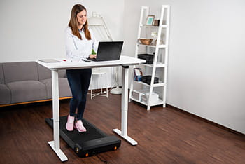 Eine Person geht in einem lockeren und eleganten Outfit mit Sportschuhen auf einem Walking Pad an einem Schreibtisch und arbeitet an einem Laptop.