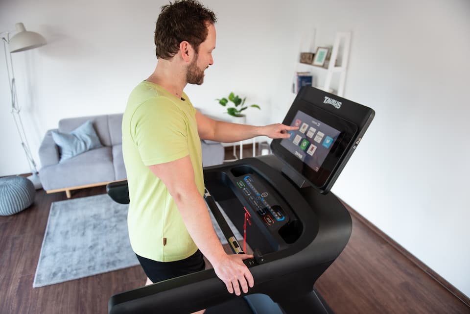 Eine Person in Sportbekleidung steht in einer wohnlichen Umgebung auf einem Laufband und tippt mit dem Finger auf das Display des Laufbands.