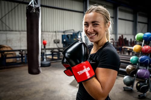 Eine Frau in einer industriellen Umgebung steht mit erhobenen Fäusten in Boxhandschuhen in der Mitte des Bildes und lächelt in die Kamera, während im Hintergrund ein Boxsack und mehrere Gewichtsbälle zu sehen sind