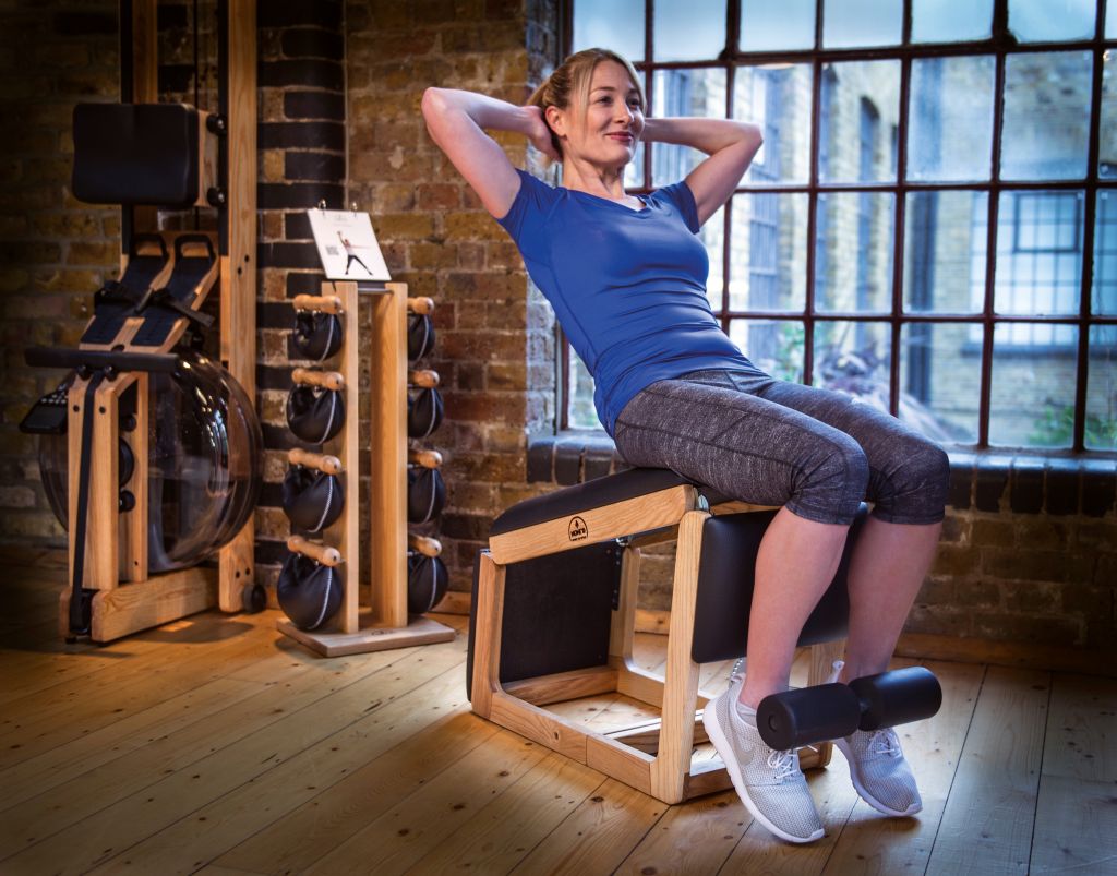 Eine Frau in Sportbekleidung sitzt auf dem TriaTrainer, ihre Beine sind an der Fußstütze eingeklemmt, da sie Sit-Ups ausführt.