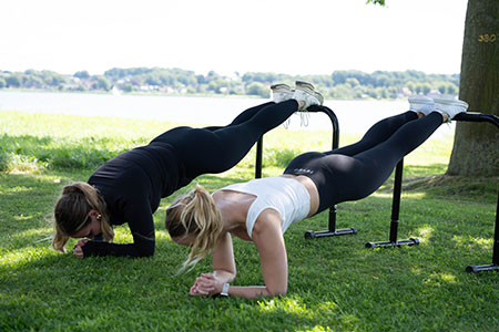 Zwei Frauen sind nebeneinander in sportlicher Kleidung und führen draußen am See eine Plank aus, ihre Füße sind dabei erhöht auf dem Multifunktionstrainer