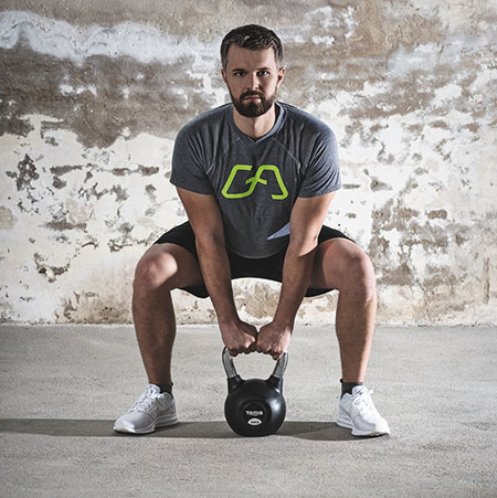 Mann in sportlicher Kleidung ist in einer niedrigen Kniebeuge-Position und hält mittig eine Kettlebell