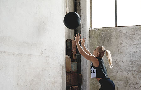 Vrouw in sportkleding staat voor een muur en gooit een medicijnbal hoog tegen de muur.