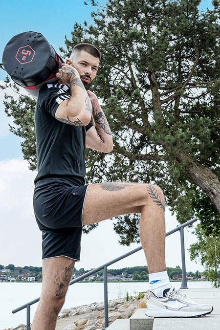 Mann in sportlicher Kleidung hat auf den Schultern einen Sandbag und geht damit im Freien Treppen hinauf.