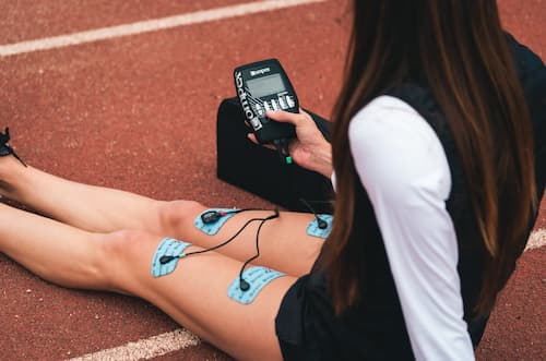 Une femme en tenue de sport assise sur une piste en tartan avec quatre électrodes d'un stimulateur musculaire sur ses cuisses, tout en contrôlant les électrodes sur l'appareil portatif