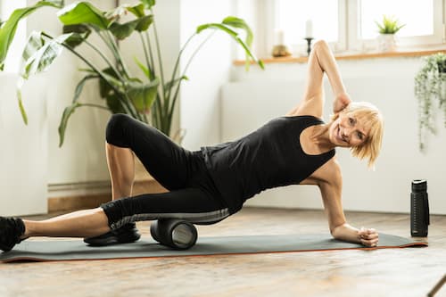 Frau in sportlicher Kleidung auf einer Yogamatte, die in einer Sideplank auf einer Faszienrolle ihren Oberschenkel massiert