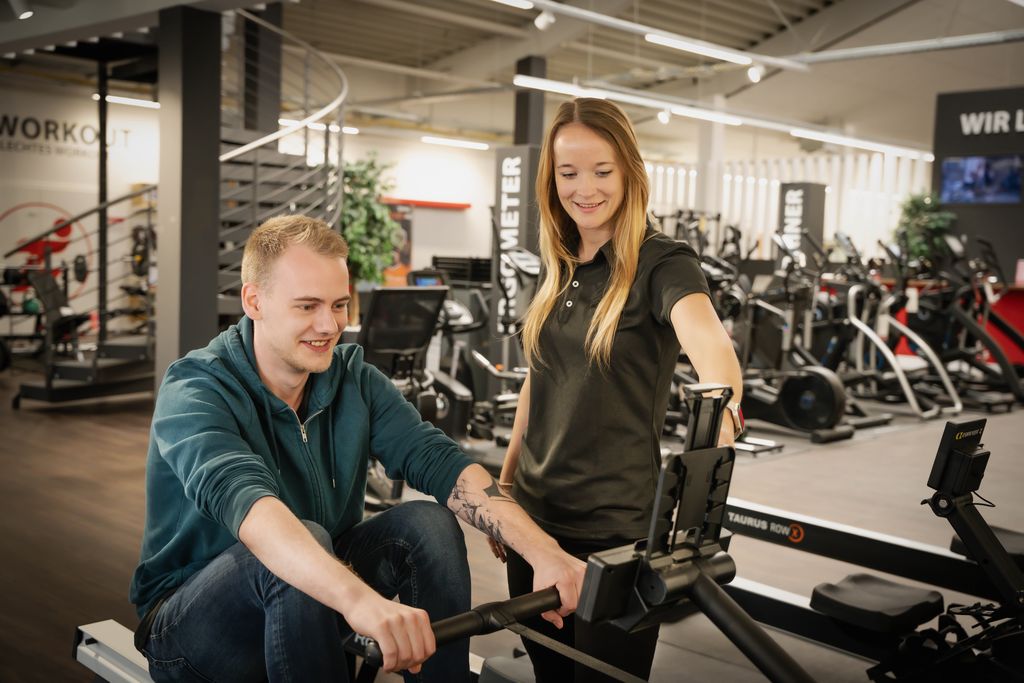 Vrouw in Fitshop-filiaal adviseert een klant die op een roeitrainer zit en deze uitprobeert.