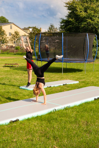 Deux filles font chacune de la gymnastique sur un tapis AirTrack, tandis qu'en arrière-plan, un enfant est sur un trampoline