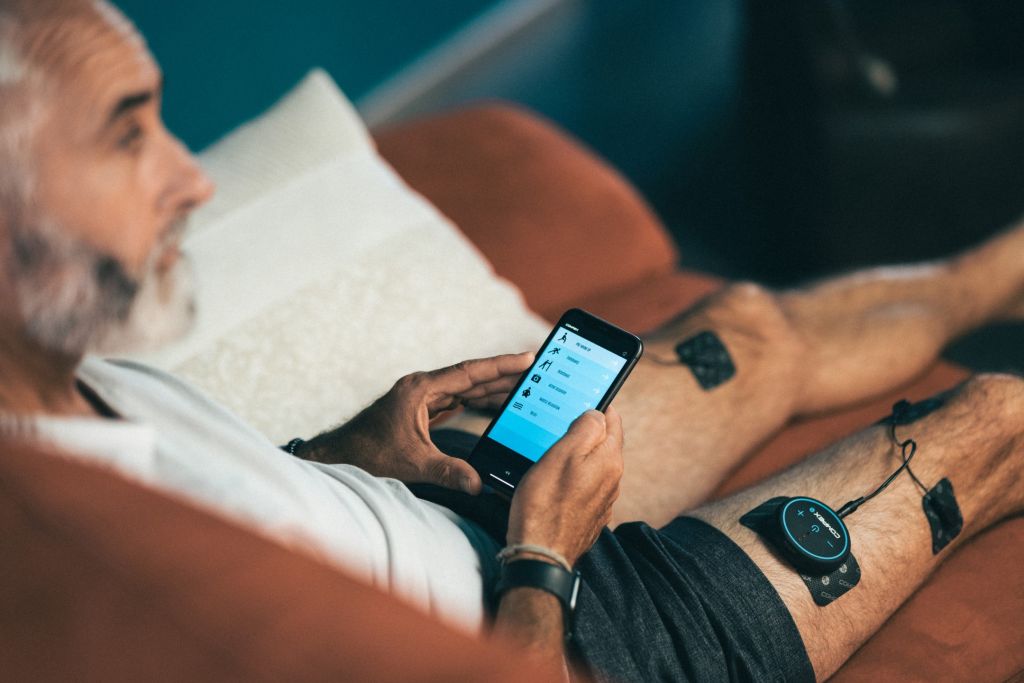 Ein Mann sitzt entspannt auf einem Sofa und hat auf den Beinen, die lang ausgestreckt sind, Elektroden kleben, die er über sein Smartphone steuert, das er in der Hand hält.