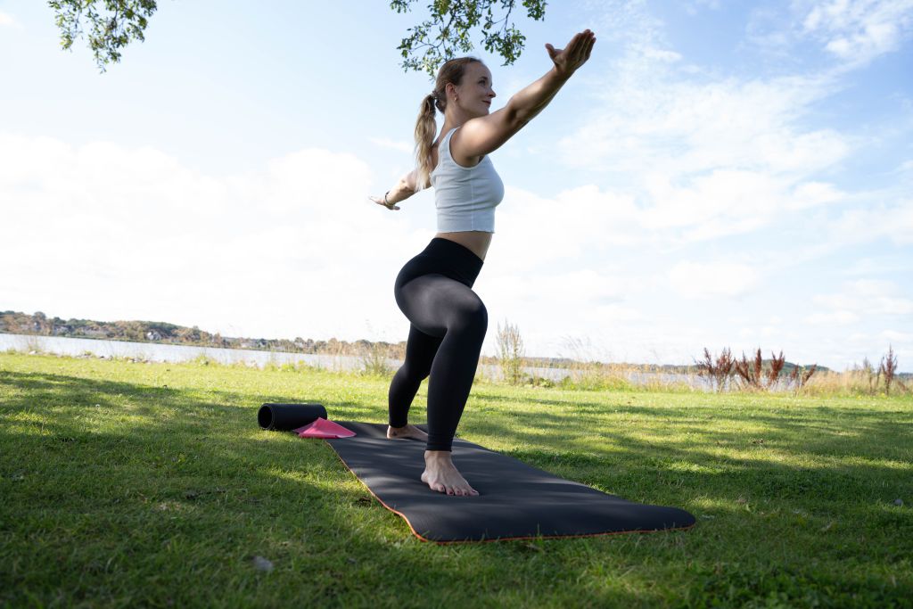 Une femme en tenue de yoga adopte la position du Guerrier II sur un tapis de yoga en l'extérieur.